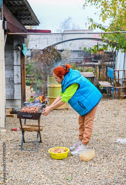 Fototapeta A 50-year-old Russian woman in a rural village grills marinated chicken outdoors, tending skewers over an open flame on a cool autumn day in Russia.
