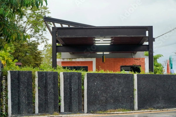 Fototapeta A modern building with a dark metal roof structure and orange accents, partially obscured by a textured black and white concrete wall and lush green foliage.