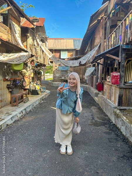 Fototapeta A cheerful young woman stands on a lively street in a traditional village, smiling brightly