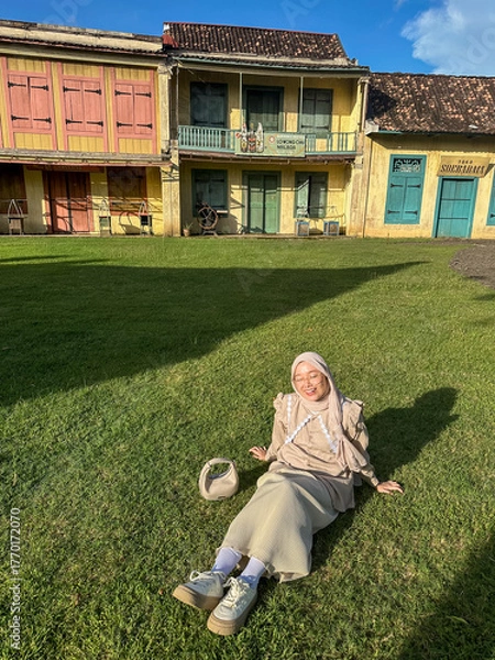 Fototapeta A person relaxes on the grass in front of a bright building under clear blue skies