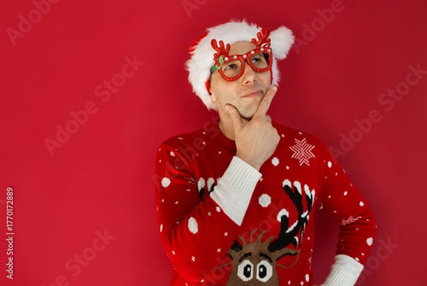 Obraz Funny man wearing a Christmas hat, showing a thinking gesture by looking up with hand on chin, contemplating an idea. Perfect for holiday-themed advertising or creative concepts.