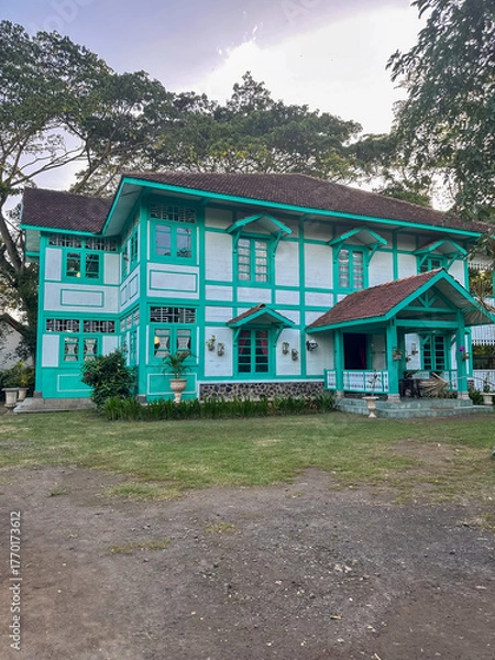 Fototapeta The colonial-style house features teal accents and a lush garden during the late afternoon