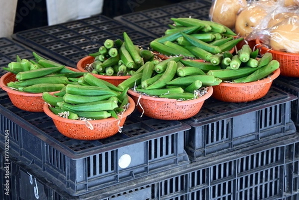 Fototapeta Five red plastic baskets filled with fresh green okra (lady's finger) on black plastic crates, light-colored round fruits visible in basket behind, vendor display at market, Taiwan.