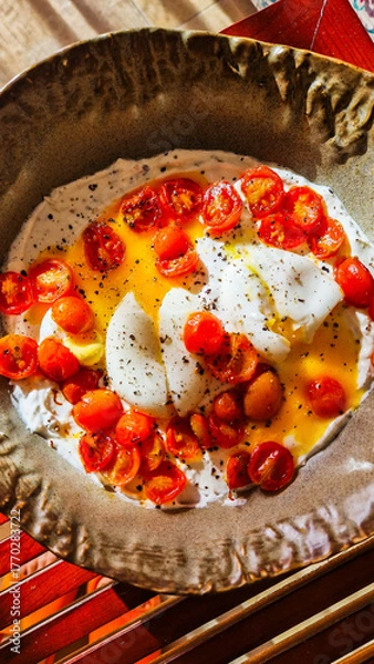 Obraz A plate of scrambled eggs topped with sliced cherry tomatoes. The dish is garnished with black pepper and served on a textured ceramic plate.