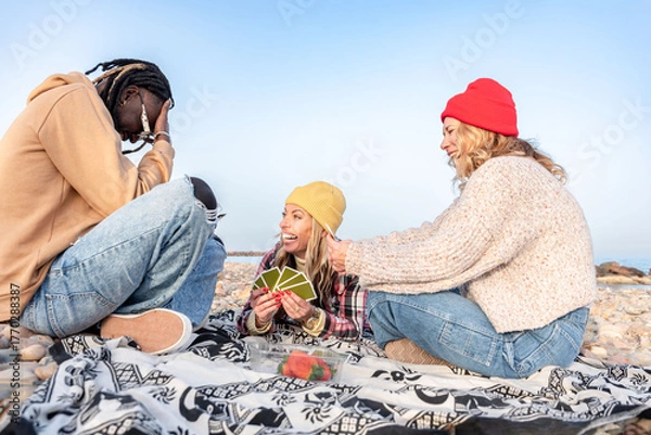 Fototapeta Group of diverse happy friends playing cards and enjoying a picnic on a rocky beach