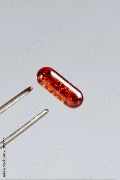 Obraz Translucent amber capsule held by tweezers on neutral background, macro pharmaceutical supplement and clinical research detail