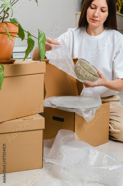 Fototapeta Portrait of woman wearing white t-shirt sitting on the floor surrounded with carton boxes and packing plates and dishes. Relocating, buying new house. Pack personal stuff from carton boxes. 