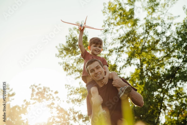 Fototapeta Father and young son enjoy bonding time in nature, with the child excitedly holding a toy airplane while sitting on dad's shoulders. Portrait of happy father with playful son outdoors. Space for text
