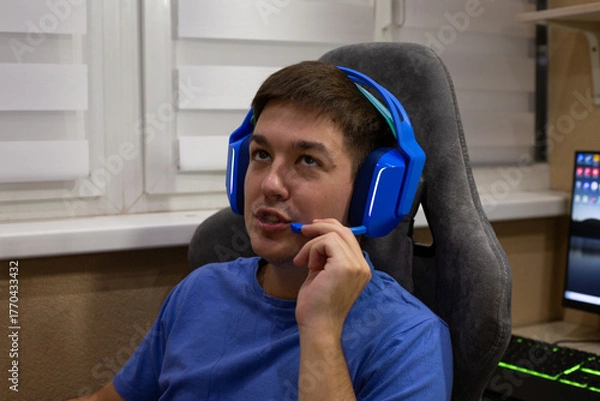 Obraz Close up of young man wearing blue gaming headphones and using microphone while sitting at gaming desk. Scene highlights gaming environment, featuring person in relaxed, focused posture