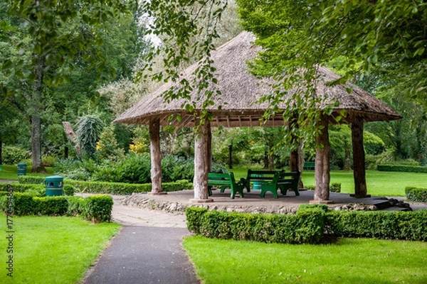 Obraz Wooden gazebo with thatched roof, located in the green area of the park. Ideal spot for outdoor relaxation and gatherings.