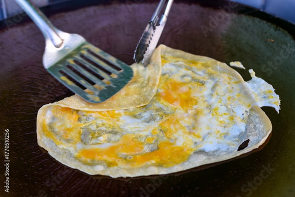Obraz An egg crepe is cooked on a hot griddle, being folded with a spatula and tongs.