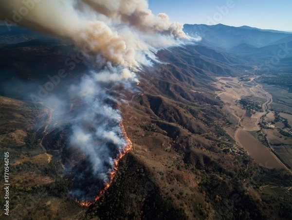 Fototapeta Aerial view of a wildfire burning through a valley covered with dense smoke and surrounding mountains