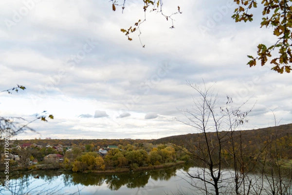 Obraz Autumn trees with yellow and red leaves on a hill above calm river reflecting forest and cloudy sky. Rural landscape photography. Autumn season and nature concept.
