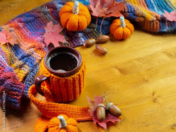Fototapeta Colorful autumn composition, a mug of hot coffee in a knitted cover on a wooden table. Knitted small pumpkins and a scarf. Copy space