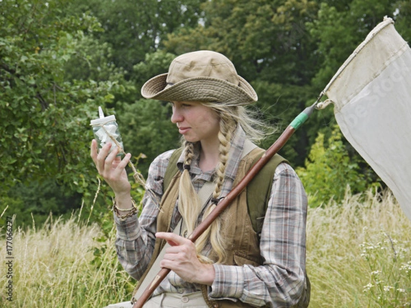 Obraz A girl entomologist in a field near a forest. Young woman with two plaits, in a hat, dressed in country style holds an insect net and a killing bottle. A student summer field period in the university.