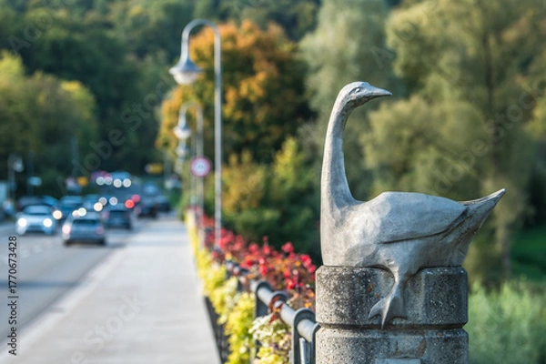 Fototapeta Skulptur einer Gans Anserinae auf der Neckarbrücke mit fließendem Verkehr in Reutlingen Mittelstadt mit Blumenschmuck am Brückengeländer. 