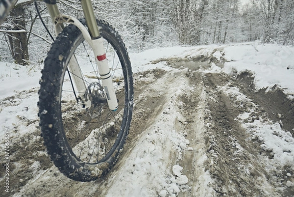 Fototapeta Bicycle wheel rides on a muddy snowy road in the forest in winter