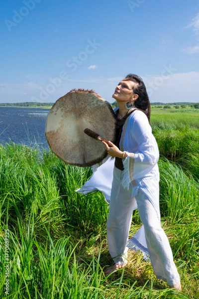 Fototapeta Middle-aged female shaman in the white dress drumming outdoors