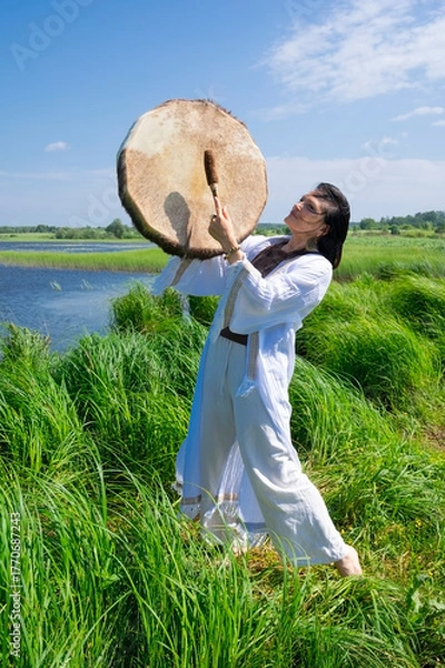 Fototapeta Female shaman in a trance in the white dress drumming in the natural environment