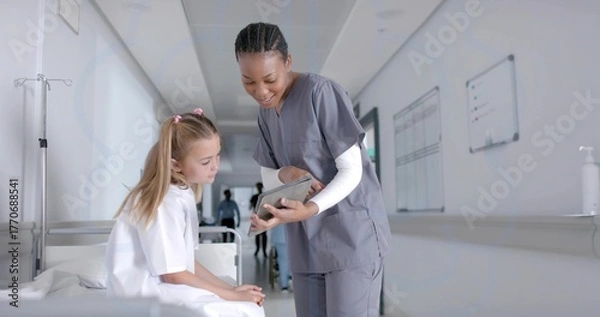 Fototapeta Leaning nurse in scrubs pointing at tablet toward child on bed in corridor, with IV stand