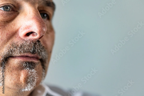 Fototapeta Detailed close-up of a middle-aged man’s lower face, showing the texture of his skin, mustache, and goatee with a neutral blue backdrop. Sad, lonely look.