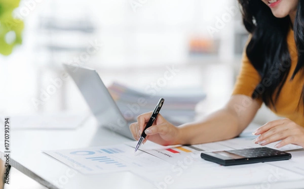 Fototapeta Focused Productivity: A woman meticulously examines financial documents, her attention fully engrossed. The modern workspace setting features a laptop and smartphone.
