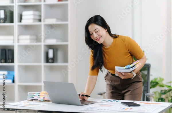 Obraz Focused Businesswoman at Work: A dedicated businesswoman, absorbed in her work, meticulously examines documents and operates a laptop.