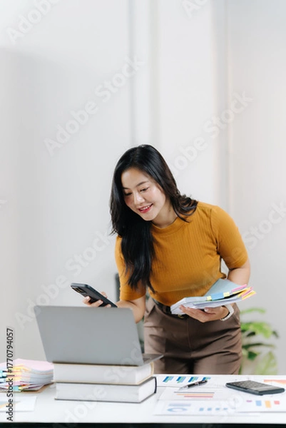 Fototapeta Efficiency in the Office: A professional woman focused in a modern workspace. She is holding swatch, and holding phone in a seamless blend of technology and design while looking towards laptop