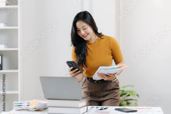 Fototapeta Digital Age Office Interaction: A focused woman in a warm-toned sweater actively engaging with her smartphone and documents, at a modern office desk setting, signifying connectivity and productivity.