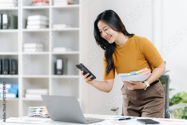 Fototapeta Focused Businesswoman At Her Desk: An Asian businesswoman, radiant and dedicated, is fully engrossed in her work at her modern workstation, with a smartphone in one hand and files in the other.