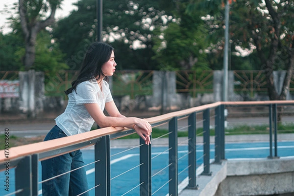 Fototapeta Young woman gazing thoughtfully outdoors, leaning on railing with trees in background during golden hour, reflecting on life and personal moments in serene park setting