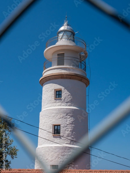 Obraz Far del Cap Gros, Port de Soller, Mallorca white lighthouse with a metal railing and a dome on top stands against a clear blue sky, viewed through a chain-link fence, Majorca