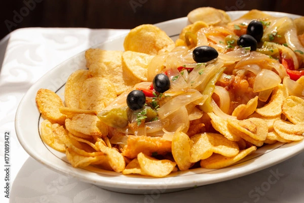 Fototapeta The "Bacalhau com Cebolada", a traditional Portuguese dish made with fried codfish topped with sautéed onions, peppers, and olives, served with crispy round potato chips.