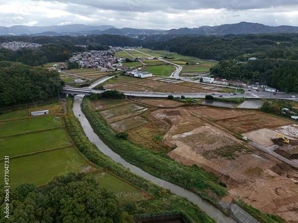 Fototapeta A rural area with a river running through it. The river is surrounded by fields and trees. The sky is cloudy and the overall mood of the image is peaceful and serene