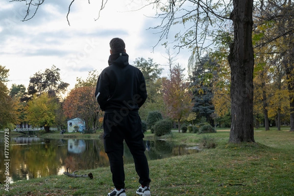 Obraz man in black hoodie and cargo pants standing by a tranquil lake in an autumn park, contemplating the scenic nature and colorful trees at sunset