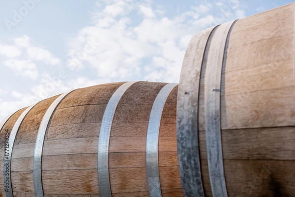 Fototapeta Close-up of oak wine barrels with metal hoops outdoors under blue sky with clouds, winery storage, traditional aging process