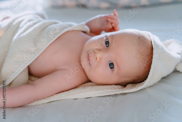 Fototapeta Happy curious baby child after bath with towel on head. Adorable little boy wrapped in blanket. Cutest 3 months old baby smiling in towel on bed close up at home. Child caring routine.