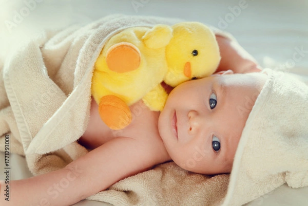 Fototapeta Happy curious baby child after bath with towel on head. Adorable little boy wrapped in blanket. Cutest 3 months old baby smiling in towel on bed close up at home. Child caring routine.