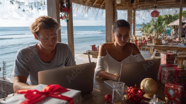 Fototapeta Christmas digital nomads working on laptops at beachside cafe with festive decorations and ocean view creating joyful holiday atmosphere