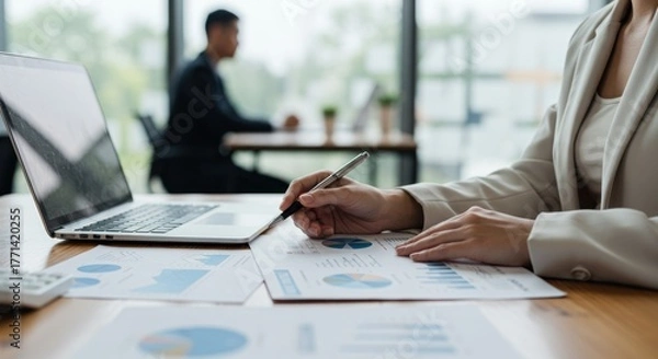 Fototapeta Businesswoman analyzes charts with laptop in office setting coworker visible in background.