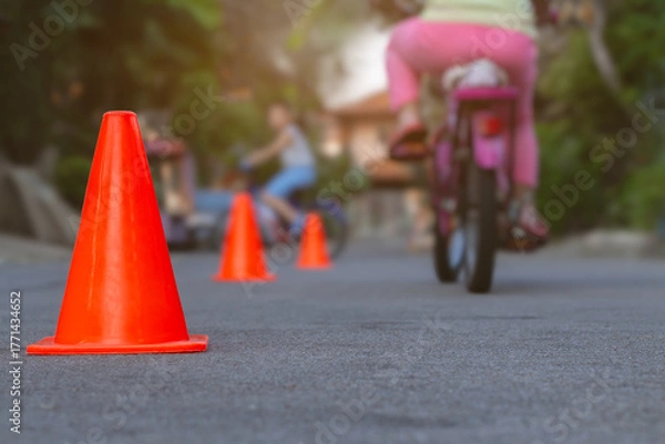Fototapeta Focus to orange traffic cones on street with blurry group of children learn to riding bicycle in the evening after back from school. Background for outdoors activity or recreation concept.