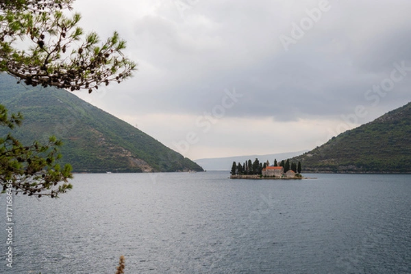 Fototapeta The small island of Saint George with its Benedictine monastery stands quietly in the Bay of Kotor, surrounded by green hills under cloudy skies.