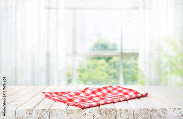 Obraz White wooden table partially covered with a red and white checkered cloth, set against a bright, blurred natural window glass