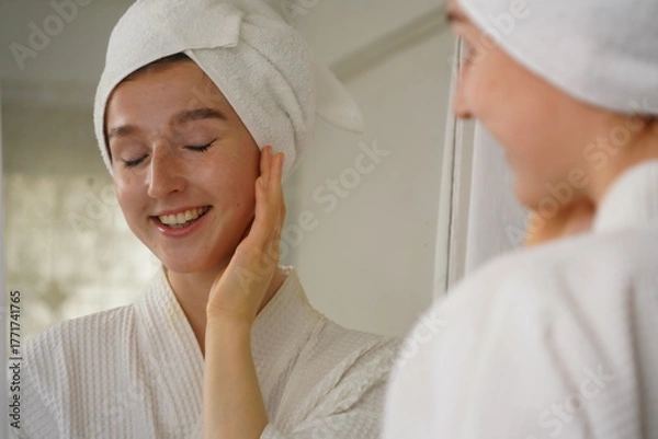 Fototapeta beautiful young woman wearing a bathrobe and using a towel to cover her hair after taking a shower looks at her face in the mirror and applies facial cream in her dressing room at home