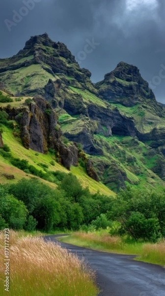 Obraz A winding road snakes through a lush valley nestled between towering, rugged mountains under a dramatic sky.