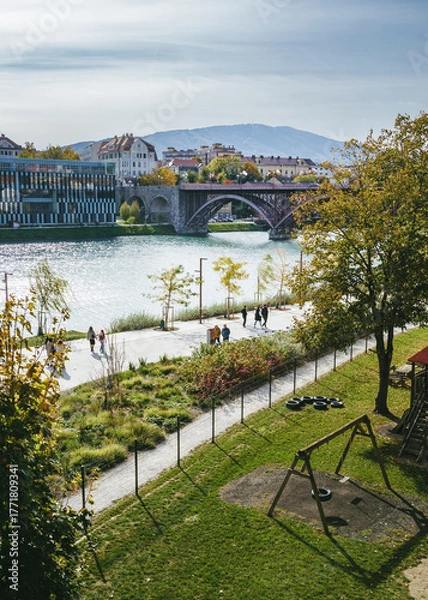 Fototapeta Cityscape of Maribor with the river Drava and the mountain Pohorje in the background. People are walking by the river towards the old bridge or stari most. There is the medicinska fakulteta