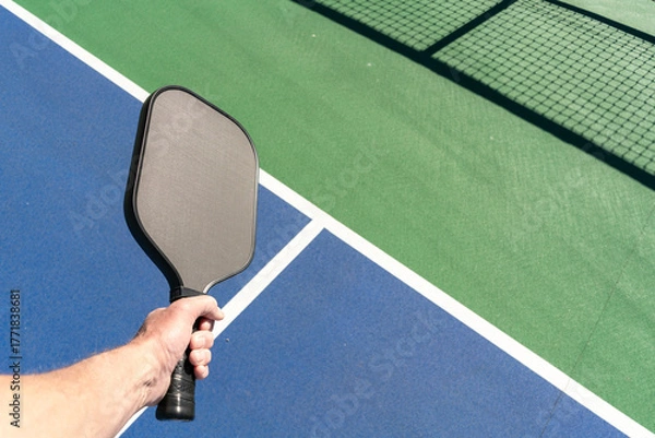 Fototapeta POV view of hand holding a pickleball paddle with a green and blue outdoor court surface in the background
