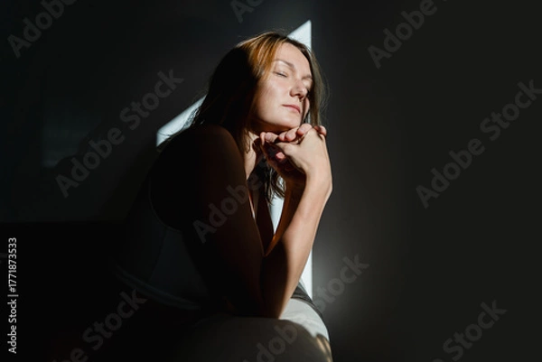 Fototapeta Deep portrait of thoughtful mature woman sitting in sunlight beam of dark room. Silhouette of sad and depressed woman at home. Concept of depression, loneliness, problems.