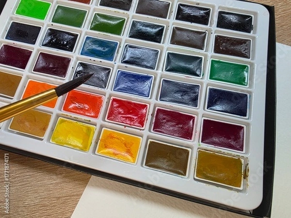 Fototapeta Watercolor paint palette with brush ready to use. Close-up of a colorful watercolor paint set with a brush, placed on a table next to blank white paper