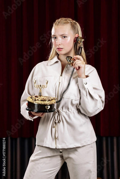Fototapeta A beautiful blonde in a beige suit holds an old desk telephone. Close-up against a theater curtain.